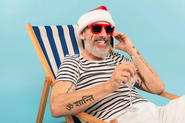 Cheerful mature man in santa hat holding glass of champagne while talking on smartphone and sitting on deck chair isolated on blue — Stock Photo