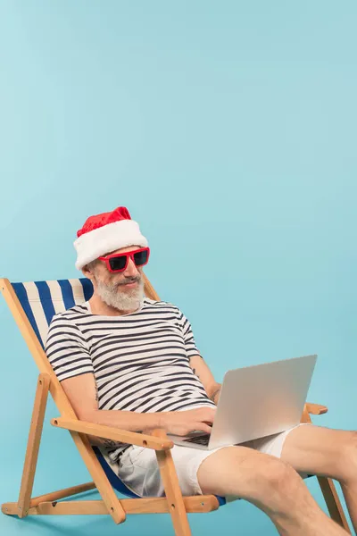 Hombre maduro feliz en sombrero de santa y gafas de sol sentado en la silla de cubierta y el uso de ordenador portátil aislado en azul - foto de stock