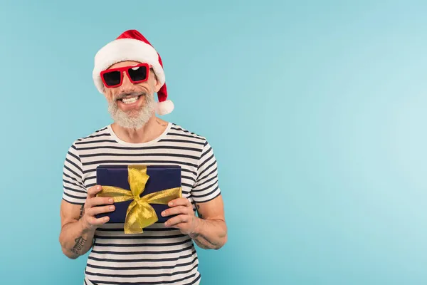 Positive middle aged man in santa hat and sunglasses holding gift box isolated on blue — Stock Photo