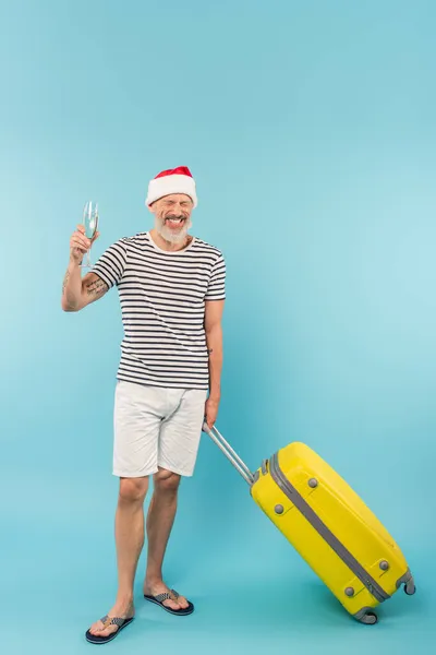 Positive middle aged man in santa hat standing with luggage and holding glass of champagne on blue — Stock Photo