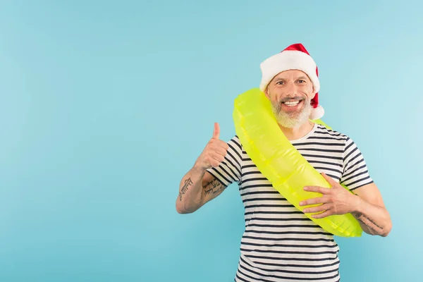 Positive middle aged man in santa hat holding inflatable ring while showing thumb up isolated on blue — Stock Photo