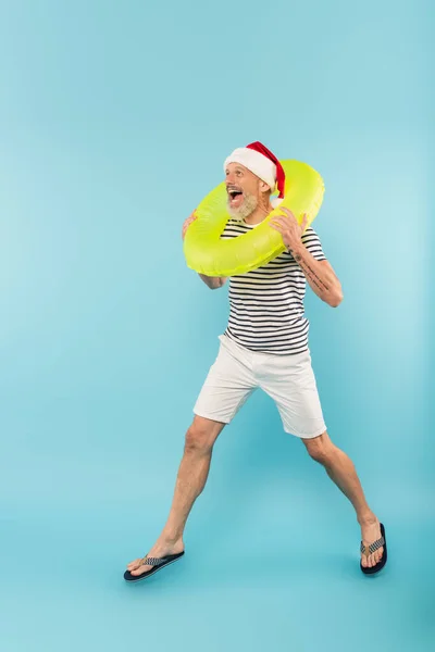 Full length of amazed middle aged man in santa hat holding inflatable ring on blue — Stock Photo