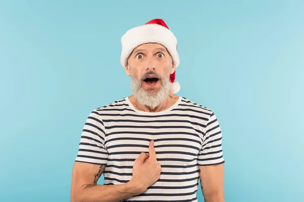 Sorprendido hombre de mediana edad en camiseta estirado y sombrero de santa señalando con el dedo a sí mismo aislado en azul - foto de stock