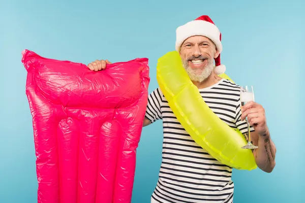 Cheerful middle aged man in santa hat and swim ring holding pink inflatable mattress and glass of champagne isolated on blue — Stock Photo