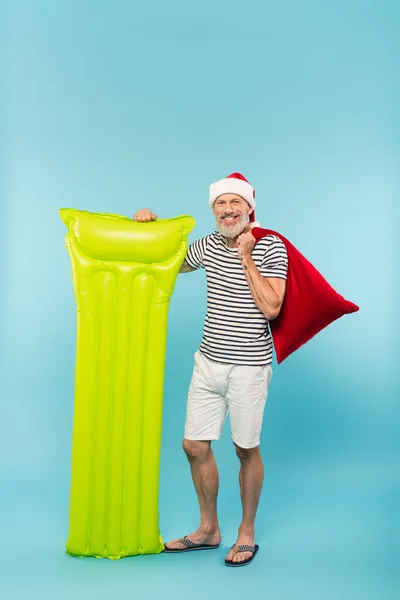 Full length of happy middle aged man in santa hat holding sack and inflatable mattress on blue — Stock Photo