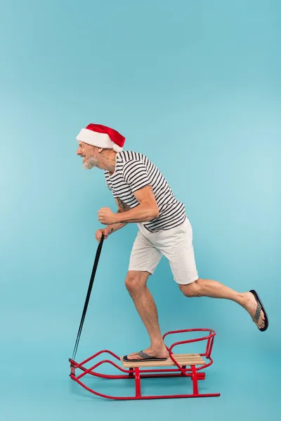 Side view of cheerful man in santa hat riding on sleigh on blue — Stock Photo