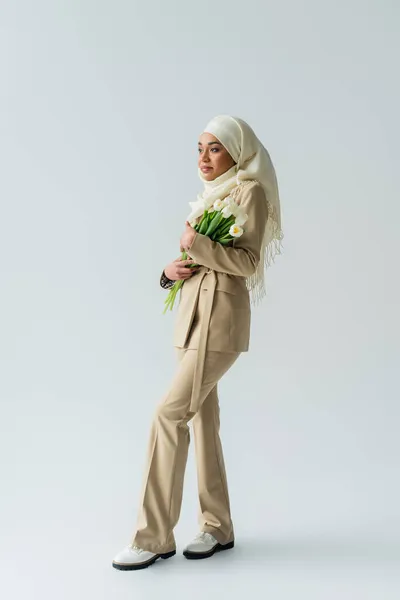 Piena lunghezza di giovane donna musulmana in hijab tenendo bouquet di tulipani su grigio — Foto stock