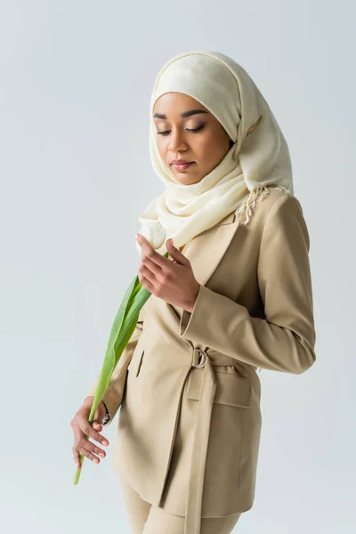 Мусульманская молодая женщина в хиджабе смотрит на тюльпан, изолированный на сером — стоковое фото