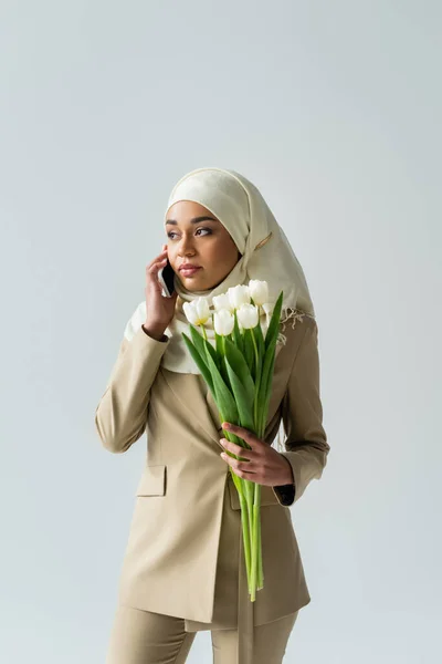 Muslimische junge Frau im Hijab hält einen Strauß Tulpen in der Hand und spricht auf dem Smartphone isoliert auf grau — Stockfoto