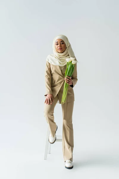 Полная длина стильной мусульманской молодой женщины в хиджабе, держащей букет тюльпанов на сером — стоковое фото