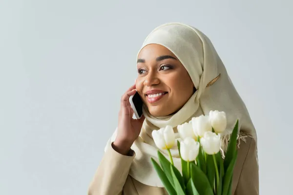 Feliz musulmana joven en hijab celebración ramo de tulipanes y hablar en el teléfono inteligente aislado en gris - foto de stock