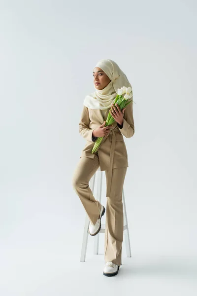 Volle Länge der stilvollen muslimischen Frau im Hijab hält Strauß von Tulpen und sitzt auf einem Schemel auf grau — Stockfoto