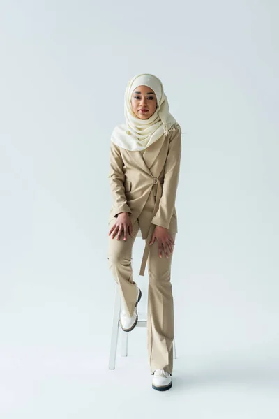 Full length of stylish muslim woman in hijab sitting on stool on grey — Stock Photo