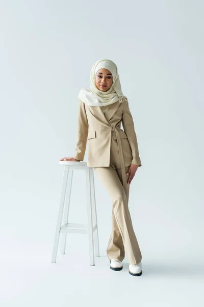 Full length of stylish muslim woman in hijab and suit leaning on stool on grey — Stock Photo