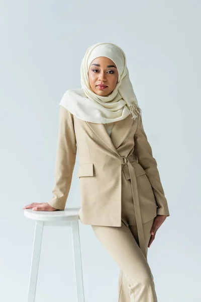 Stylish muslim woman in hijab and suit leaning on stool isolated on grey — Stock Photo