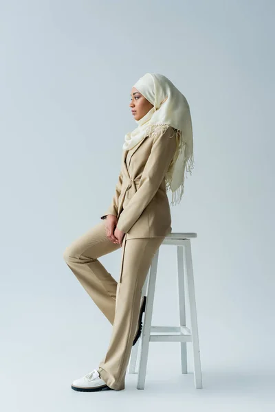 Vista lateral de la mujer musulmana con estilo en hijab y traje apoyado en el taburete en gris - foto de stock