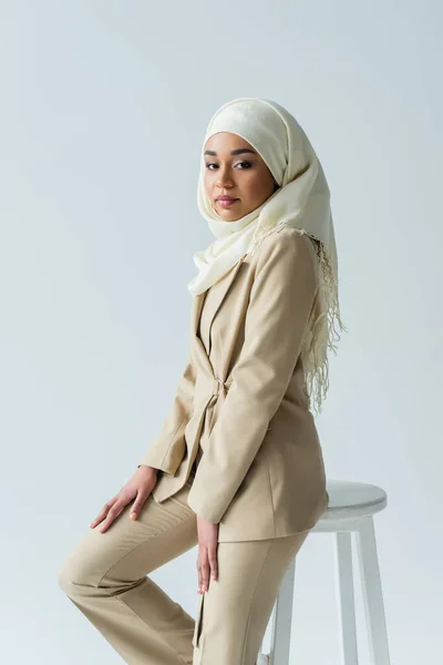 Mujer musulmana con estilo en traje hijab y beige apoyada en el taburete aislado en gris - foto de stock