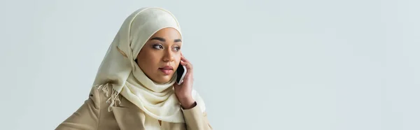 Стильная мусульманка в хиджабе и бежевом костюме разговаривает на смартфоне, изолированном на сером, баннере — стоковое фото