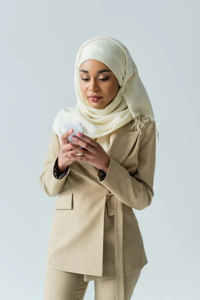 Mujer musulmana con estilo en traje hijab y beige usando teléfono inteligente aislado en gris - foto de stock
