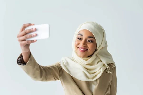 Mujer musulmana feliz en traje hijab y beige tomando selfie en el teléfono inteligente aislado en gris - foto de stock