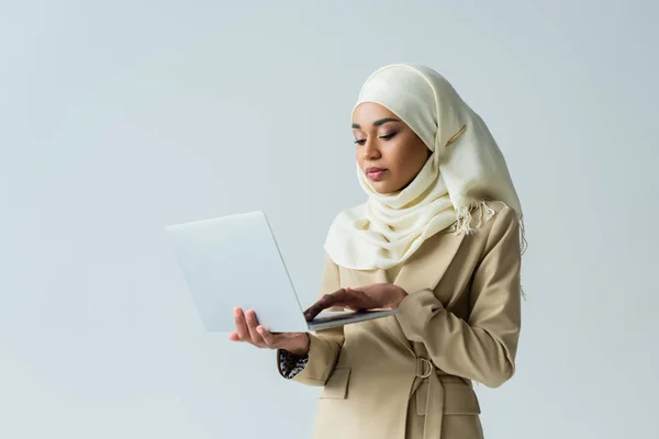 Стильная мусульманка в хиджабе и бежевом костюме, печатающая на ноутбуке, изолированном на сером — стоковое фото