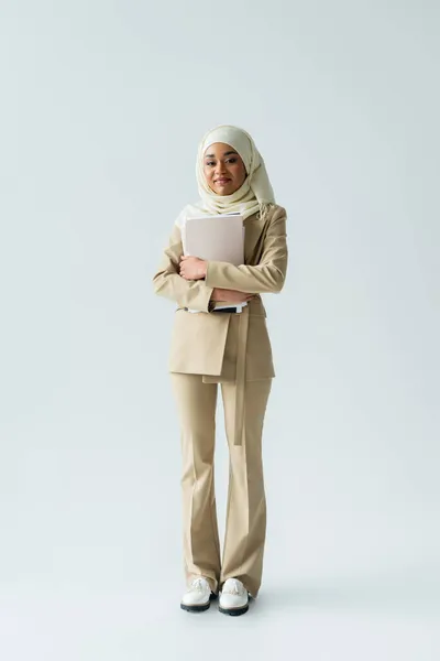 Full length of happy muslim woman in hijab and beige suit holding folders on grey — Stock Photo