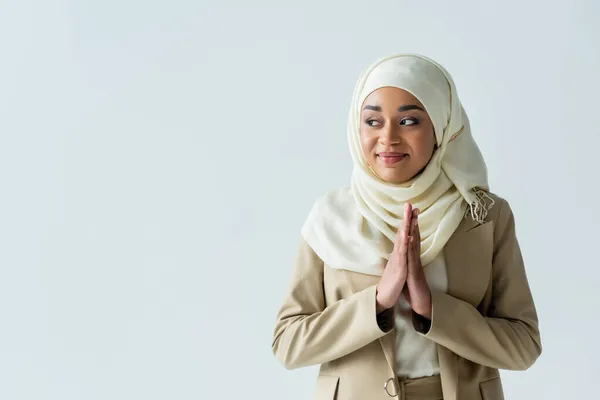 Feliz mujer musulmana en hijab de pie con las manos orantes y sonriendo aislado en gris - foto de stock