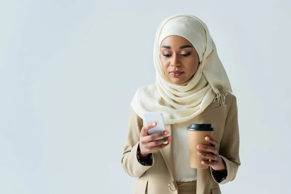 Jeune femme musulmane en hijab tenant une tasse en papier et utilisant un smartphone isolé sur gris — Photo de stock