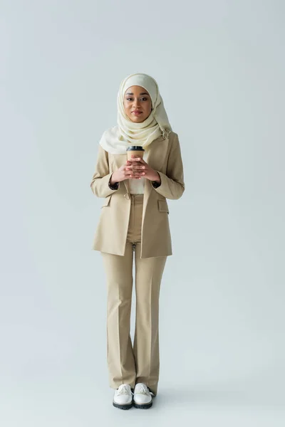 Full length of young muslim woman in hijab holding paper cup on grey — Stock Photo