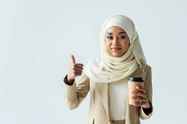 Happy muslim woman in hijab holding paper cup and showing thumb up isolated on grey — Stock Photo