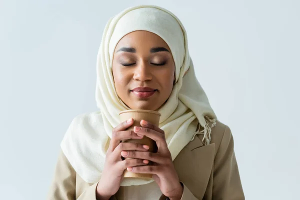 Молодая мусульманка в хиджабе держит бумажную чашку и нюхает кофе, чтобы изолироваться на сером — стоковое фото