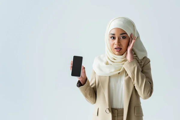 Шокированная мусульманка в хиджабе держит смартфон с пустым экраном, изолированным на сером — стоковое фото