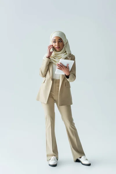 Longitud completa de la joven musulmana en hijab sosteniendo la tableta digital y hablando en el teléfono inteligente en gris - foto de stock