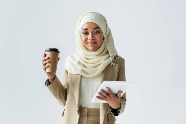 Alegre mujer musulmana en hijab sosteniendo tableta digital y café para ir aislado en gris - foto de stock