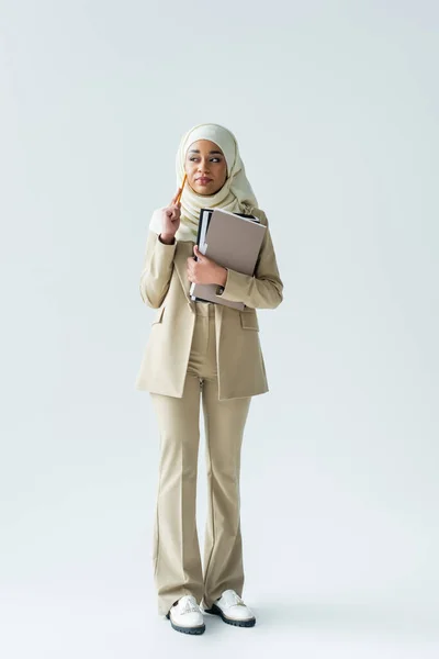 Full length of pensive muslim woman in hijab holding pencil and folders on grey — Stock Photo