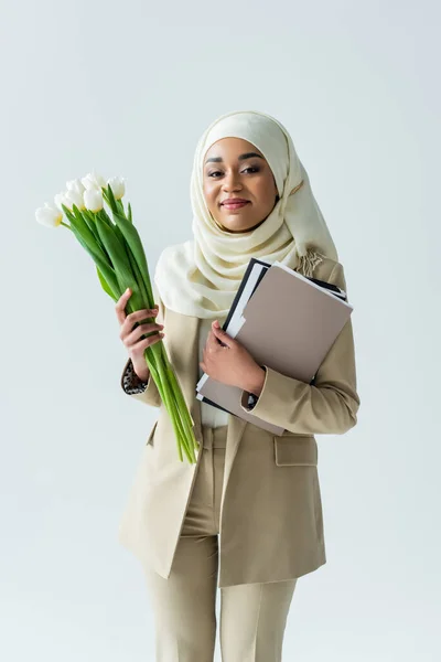 Happy muslim businesswoman in hijab holding tulips and folders isolated on grey — Stock Photo