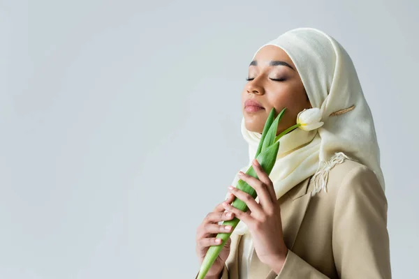 Молода мусульманка з закритими очима тримає тюльпан ізольовано на сірому — стокове фото