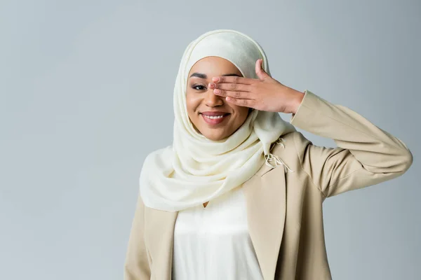 Retrato de mujer musulmana feliz en hijab que cubre el ojo mientras posa aislado en gris - foto de stock
