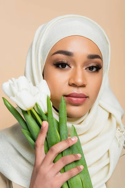 Portrait of pretty muslim woman in hijab with makeup holding tulips isolated on beige — Stock Photo