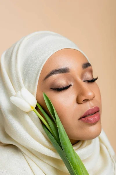 Porträt einer hübschen muslimischen Frau im Hijab mit geschlossenen Augen in der Nähe von Tulpe isoliert auf beige — Stockfoto