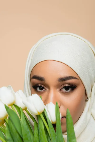 Retrato de mujer musulmana en hijab mirando a la cámara a través de tulipanes aislados en beige - foto de stock