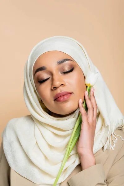 Portrait of young muslim woman in hijab with closed eyes holding tulip isolated on beige — Stock Photo