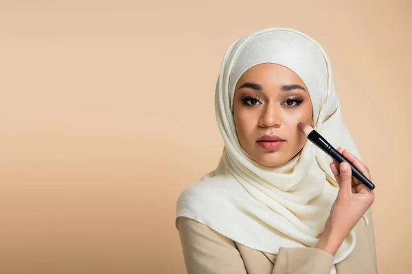 Jovem mulher muçulmana no hijab aplicando maquiagem fundação isolada no bege — Fotografia de Stock