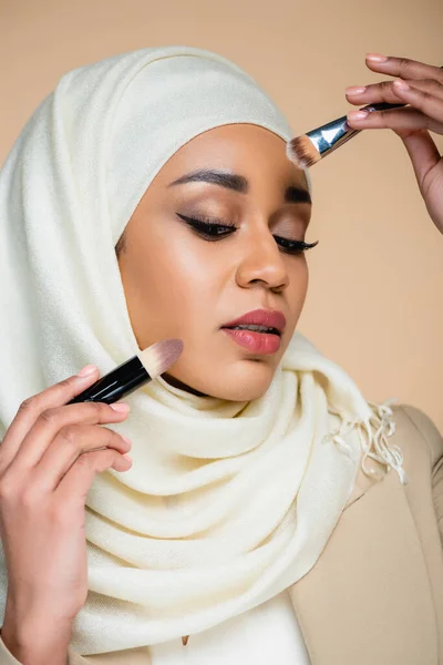 Joven mujer musulmana en hijab sosteniendo cepillos cosméticos aislados en beige - foto de stock