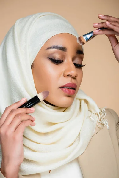Young muslim woman in hijab holding cosmetic brushes isolated on beige — Stock Photo