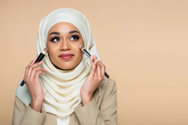 Smiling muslim woman in hijab applying face foundation with cosmetic brushes isolated on beige — Stock Photo