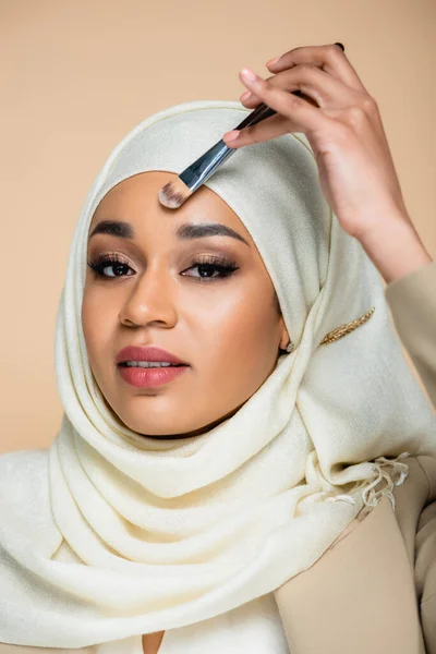 Junge Muslimin im Hijab trägt Gesichtsfundament mit Kosmetikbürste isoliert auf beige auf — Stockfoto