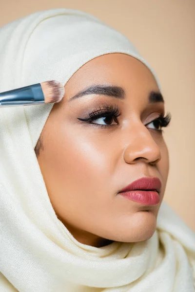 Retrato de mujer joven musulmana en hijab aplicando base facial con cepillo cosmético aislado en beige - foto de stock