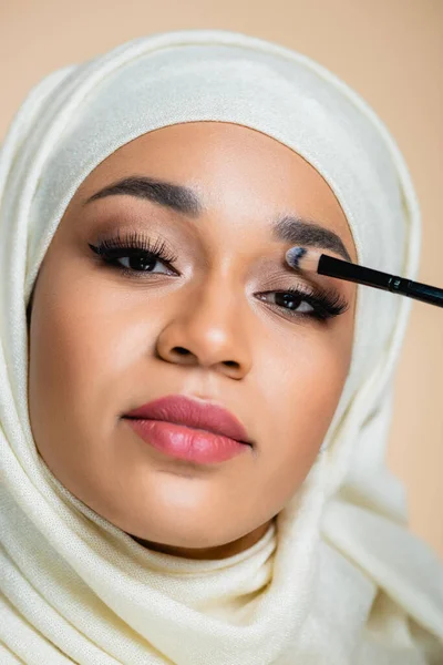 Primer plano de la joven musulmana en hijab aplicando sombra de ojos aislada en beige - foto de stock