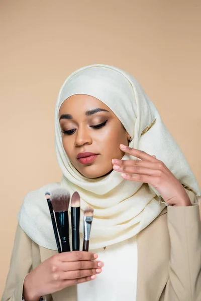 Junge muslimische Frau im Hijab betrachtet eine Reihe von Kosmetikbürsten isoliert auf beige — Stockfoto